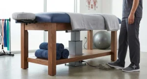 An electric stationary massage table with a foot pedal control showing the hydraulic lift mechanism for adjusting height in a physical therapy clinic.